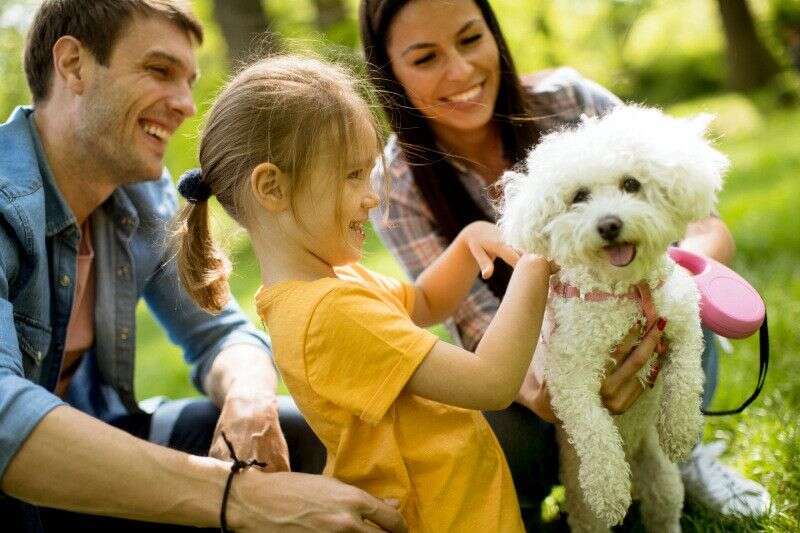 A cheerful family is sitting on grass, smiling while a young girl pets a fluffy white dog.