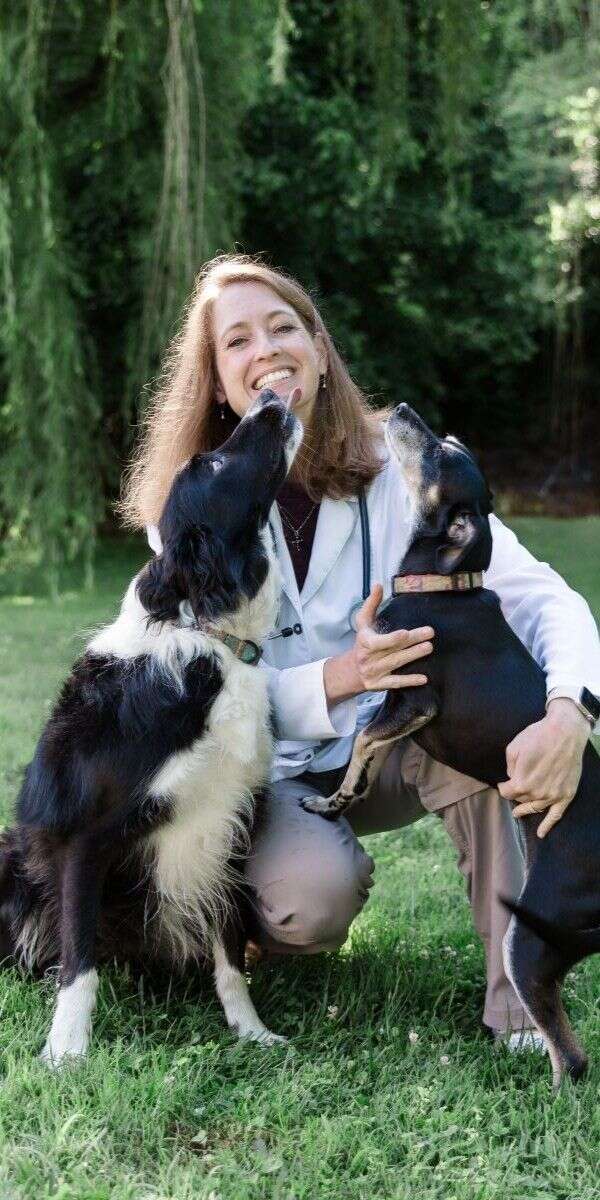 Person kneeling on grass, smiling and holding two dogs that are looking up at them.